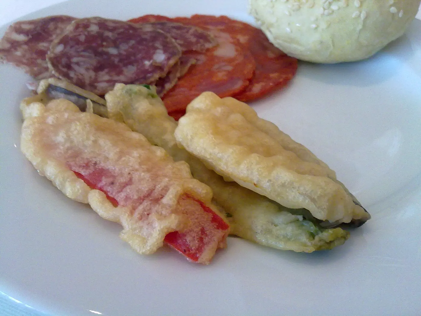 Battered and fried green beans and red peppers in the foreground of a white plate, with slices of salami and a bread roll behind