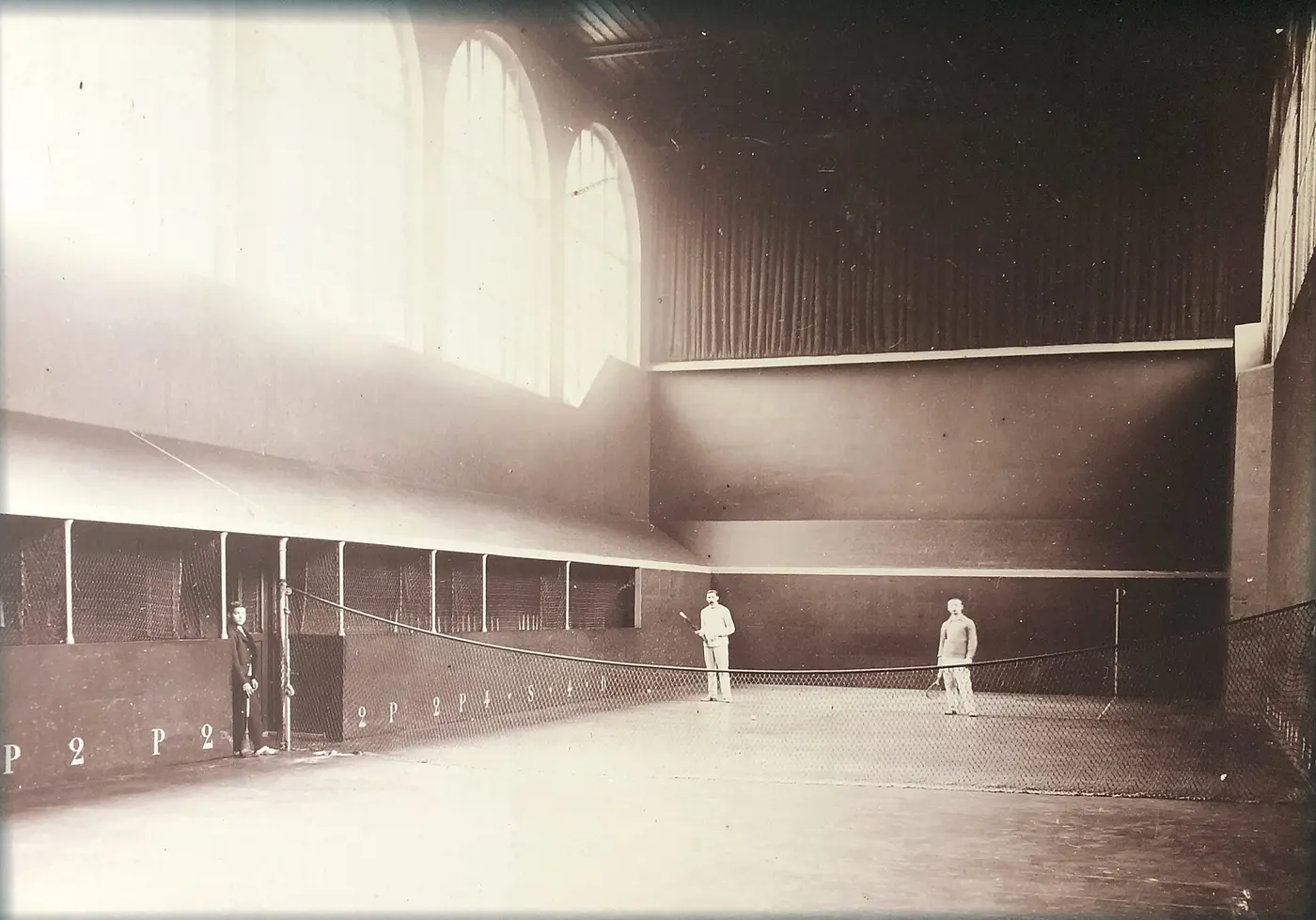 Jeu de paume players standing on either side of a net on an indoor court enclosed by high walls