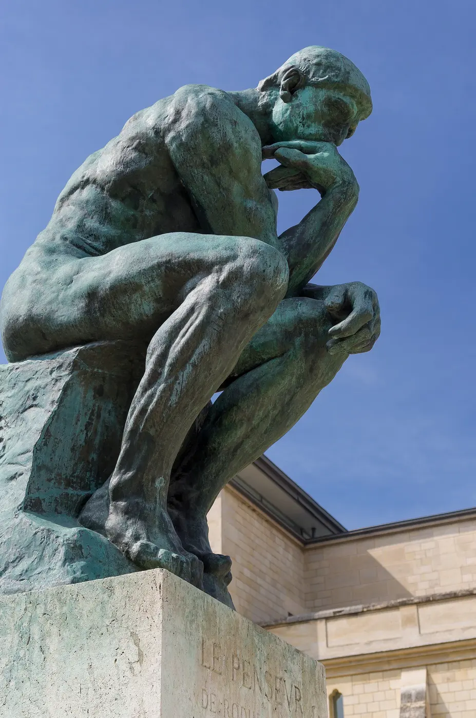 A bronze statue of a naked man on a pedestal, seated with his right elbow on his left thigh and his chin on his hand