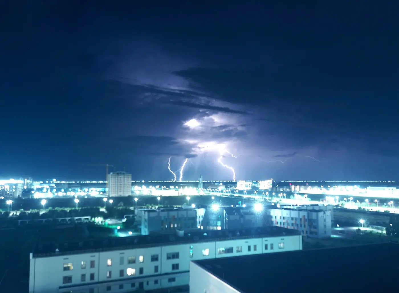 Several bolts of lightning flash beneath distant clouds beyond the lights of a city at night
