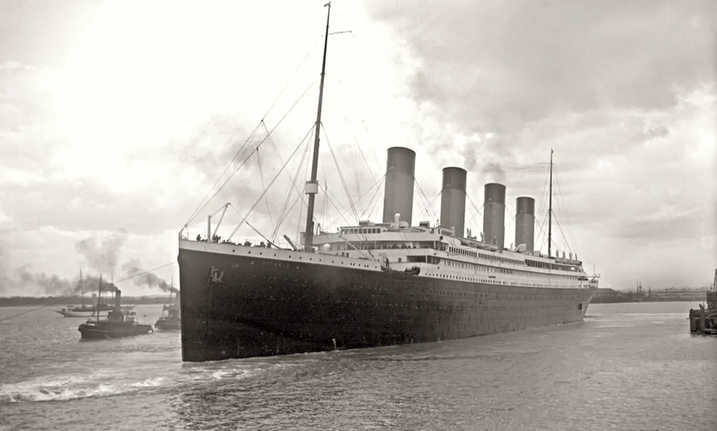 A black-and-white photo of a large passenger liner with four funnels leaving port, surrounded by small boats