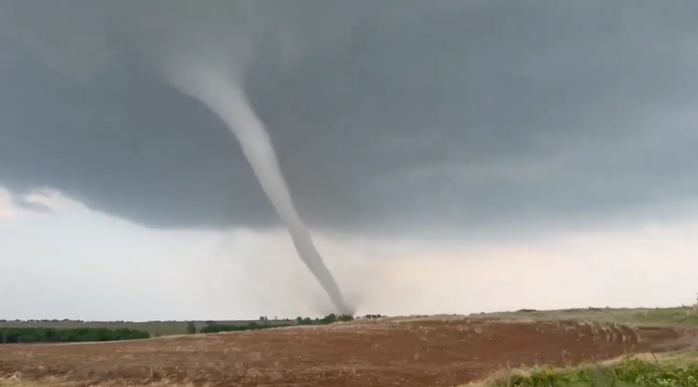 A slender tornado reaching from the base of a dark cloud down to the ground beyond a wide field