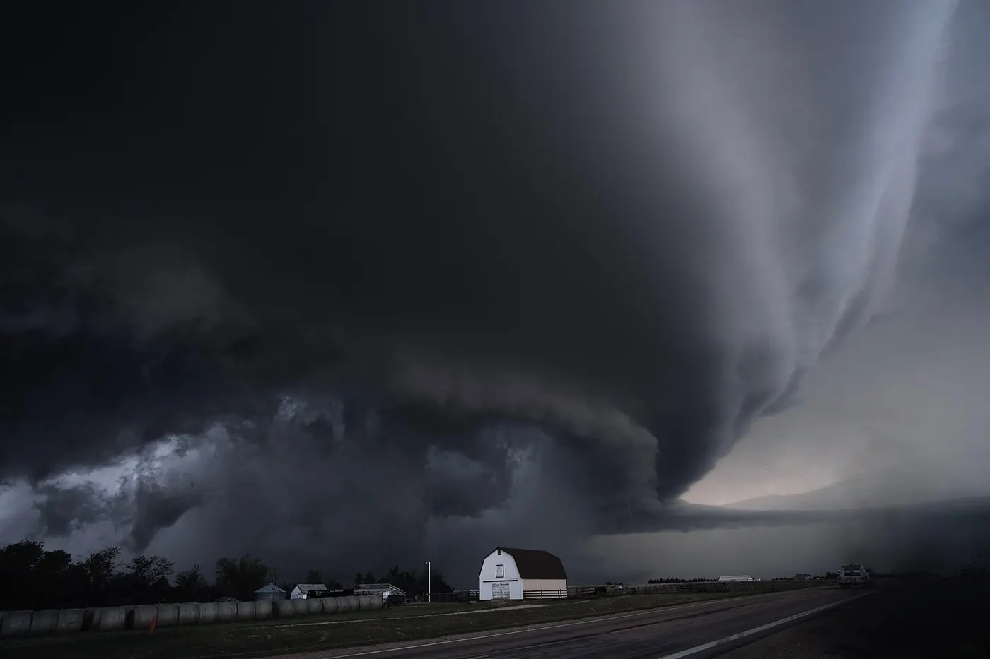 A huge, dark thunderstorm with a swirling shape spreading beyond a farm barn