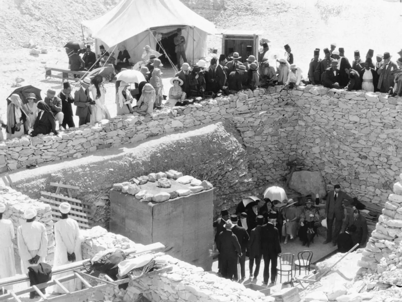 Black-and-white photo of crowds in hats and with parasols gathered around a stone-walled tomb entrance