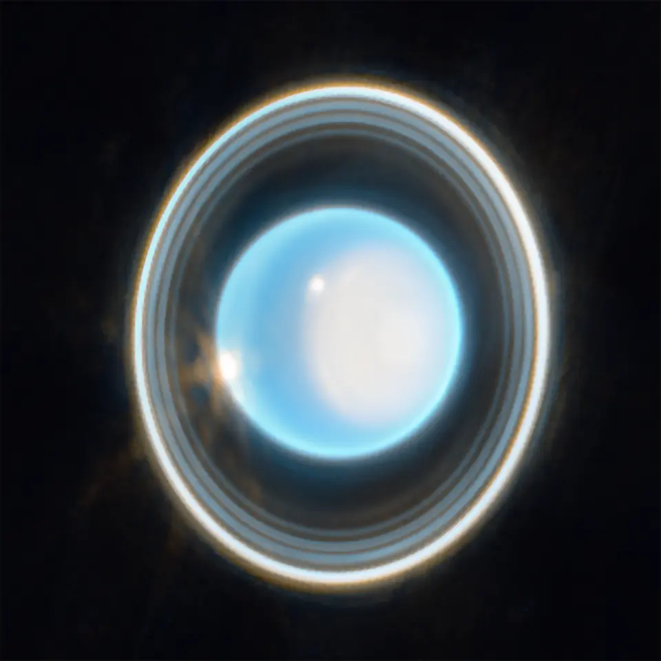 The pale blue globe of Uranus encircled by several thin rings, with a bright white glow near the pole on the right
