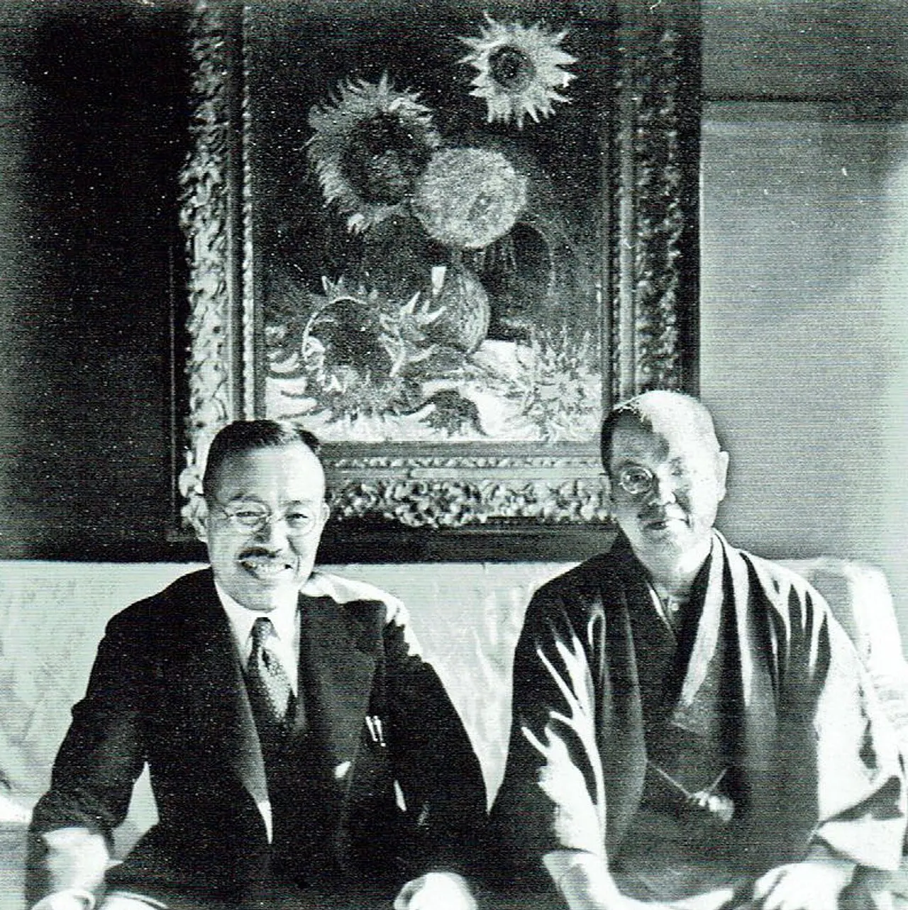 A black-and-white photo of a man in a suit and a man in a kimono seated side by side on a sofa in front of a framed painting of sunflowers