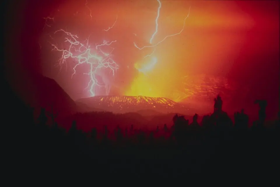 Several branching bolts of lightning flash through a red-lit eruption plume, with trees silhouetted in the foreground