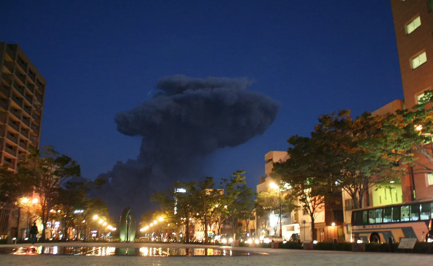 Beyond a city plaza at dusk, a black plume of ash rises high like a mushroom