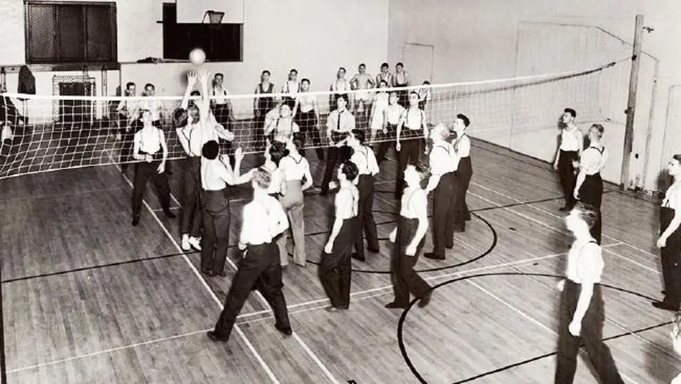 Black-and-white photo of a large group of men in shirts and trousers hitting a ball over a net stretched across a gym