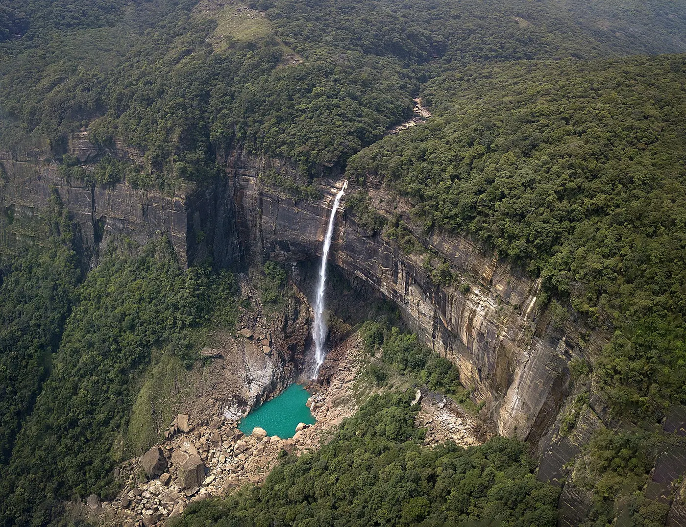 A single ribbon of waterfall dropping from a high, forest-covered cliff into a pool of blue-green water