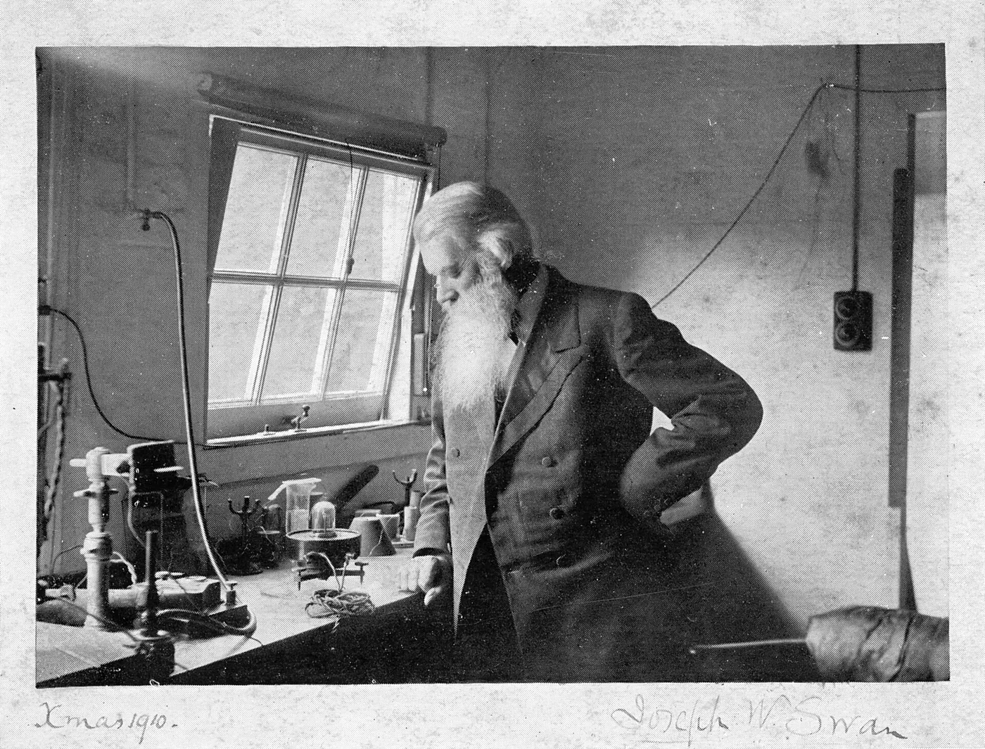 A black-and-white photo of a man with a long white beard standing at a desk covered in laboratory equipment