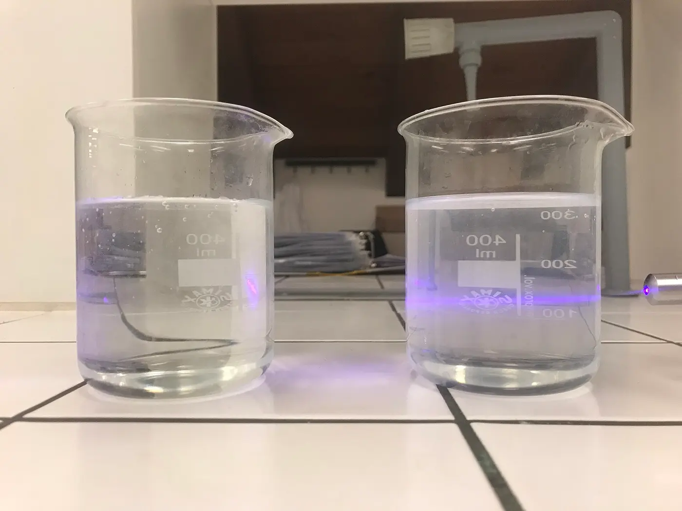 Two beakers lit from the side, one with water and one with water mixed with a little milk; a beam of light is visible only in the right-hand beaker