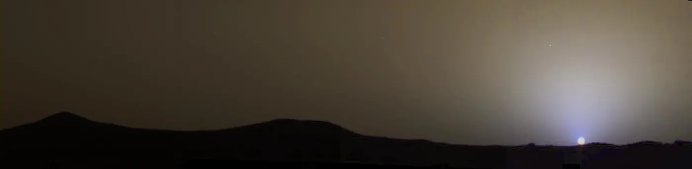 Above a line of dark hills, in a brownish sky, only the area around the setting Sun glows bluish white