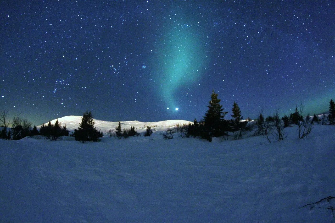 Two especially bright points of light side by side in a starry sky above snow-covered hills and trees, with a faint green aurora