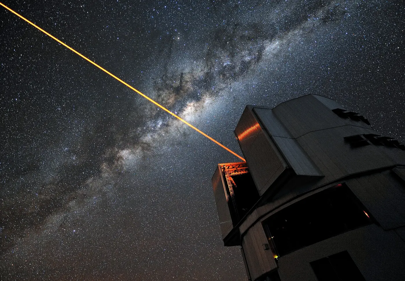 An orange laser beam shooting straight from a telescope dome into a starry sky crossed by the Milky Way