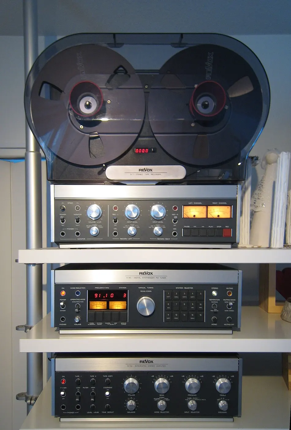 A reel-to-reel tape recorder with two large reels stacked on shelves above a tuner and an amplifier lined with knobs