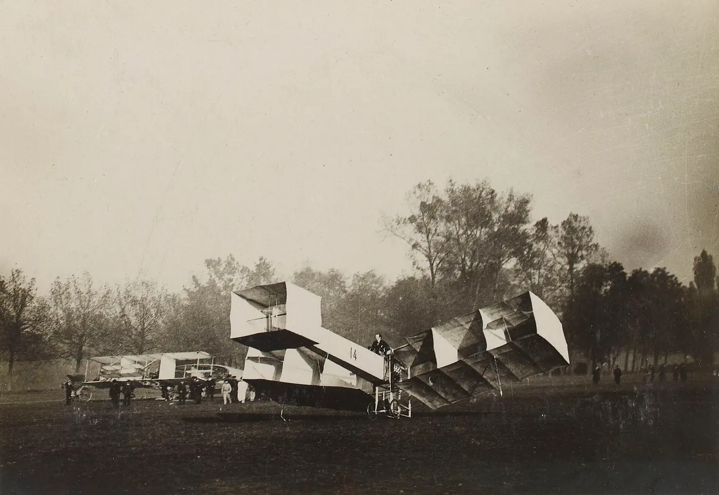 Black-and-white photo of an early airplane built from box-shaped wings, with people standing around it