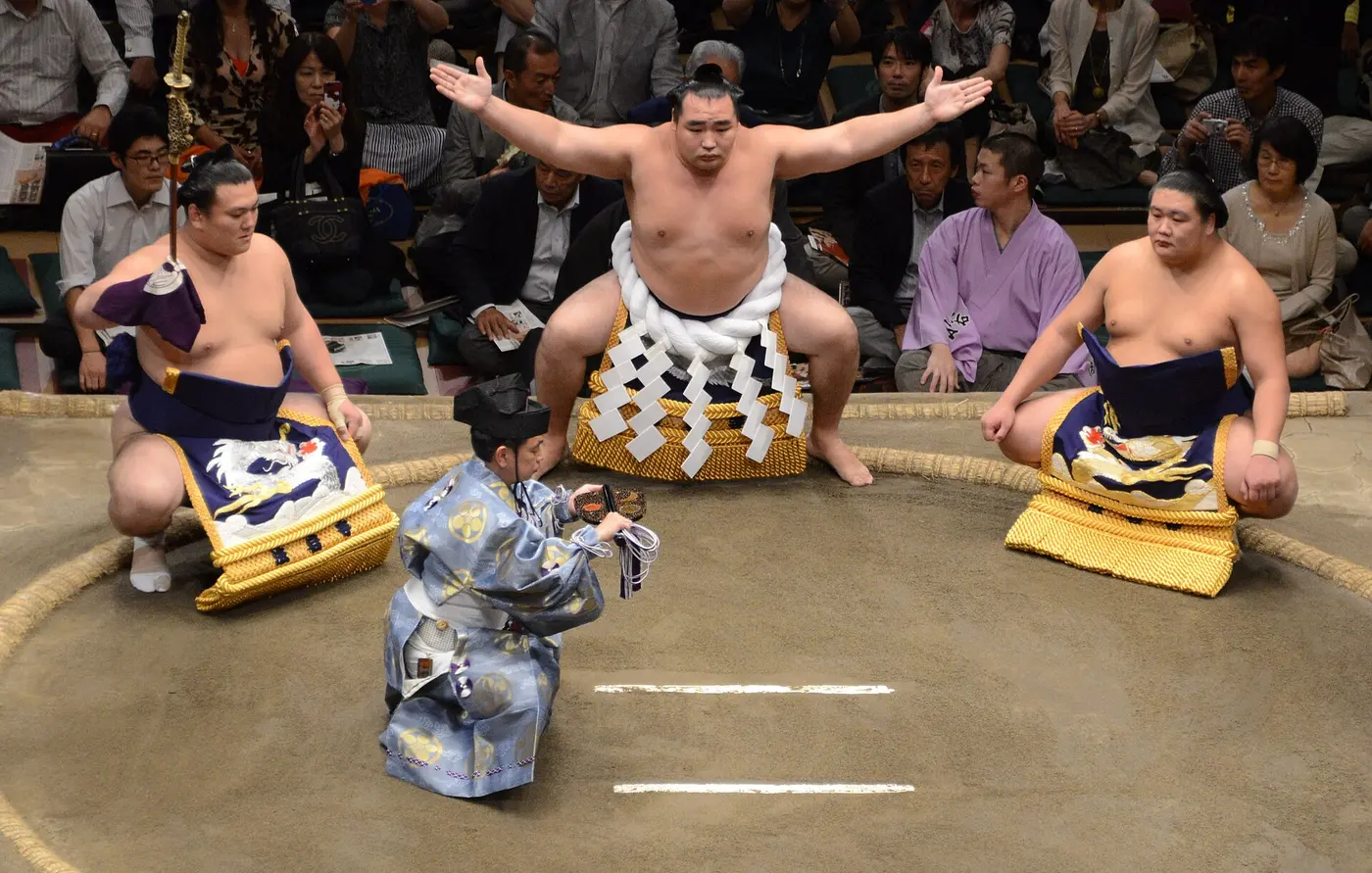 A yokozuna wearing a thick white rope spreads his arms wide, with a wrestler crouching on either side